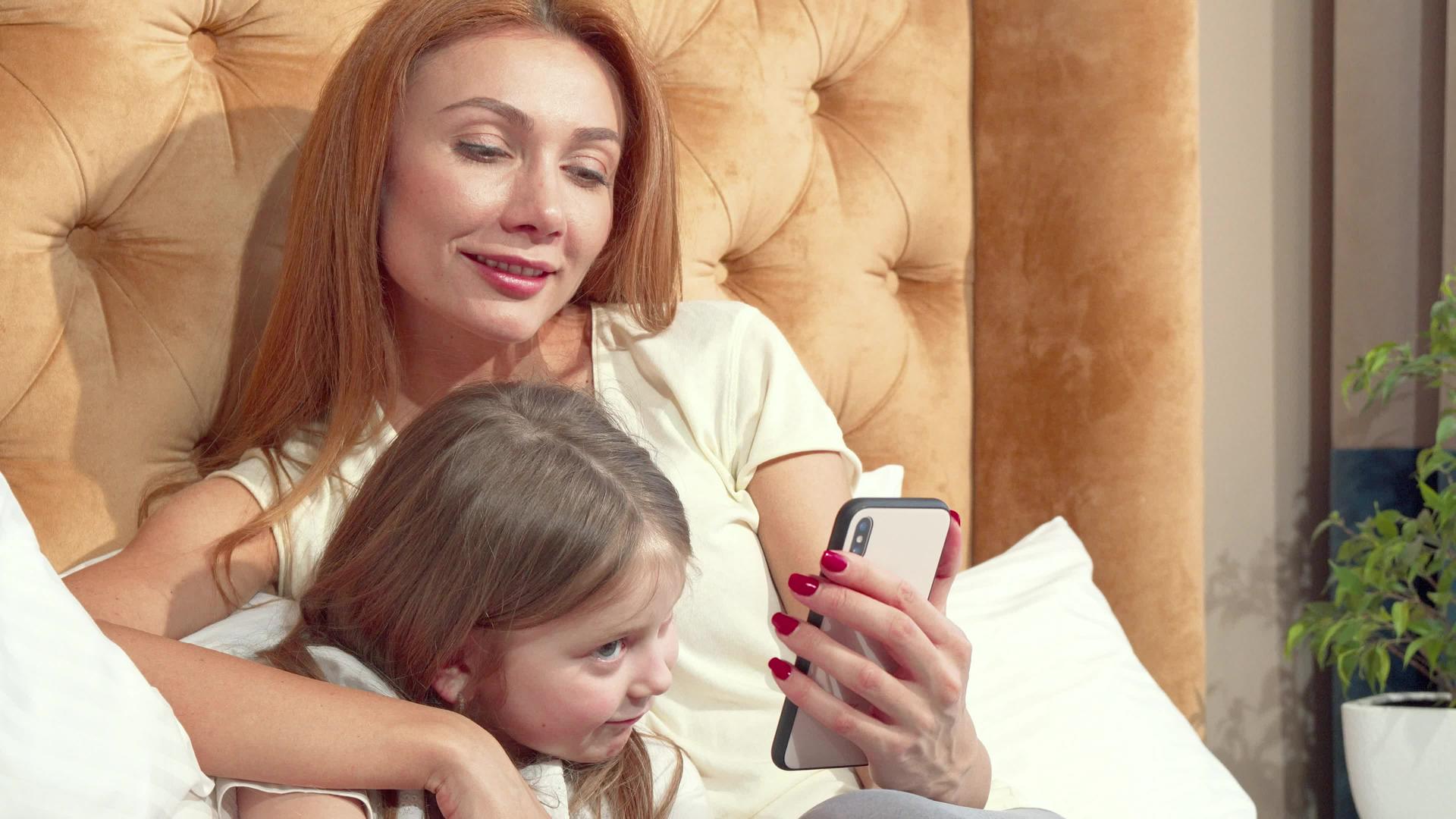 美丽的女人和可爱的小女儿在家里用智能手机玩耍视频的预览图