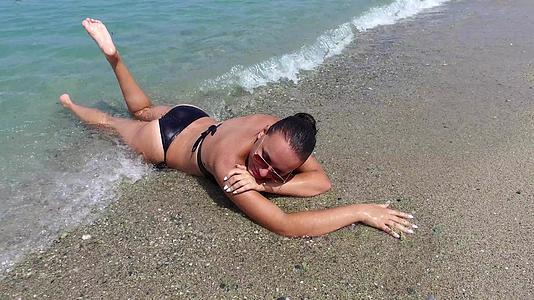 一个穿着黑色泳衣的性感年轻女子躺在海滩上被海浪喷洒视频的预览图