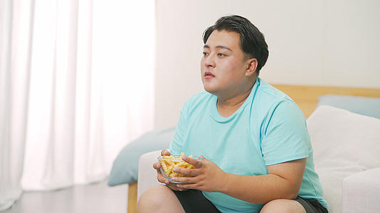 4k胖男生坐在沙发前边吃垃圾食品边看电视视频的预览图