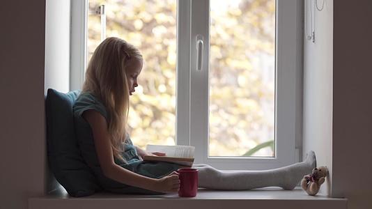 穿着绿色裙子的女孩在窗台上读书喝红杯泰迪熊的饮料视频的预览图