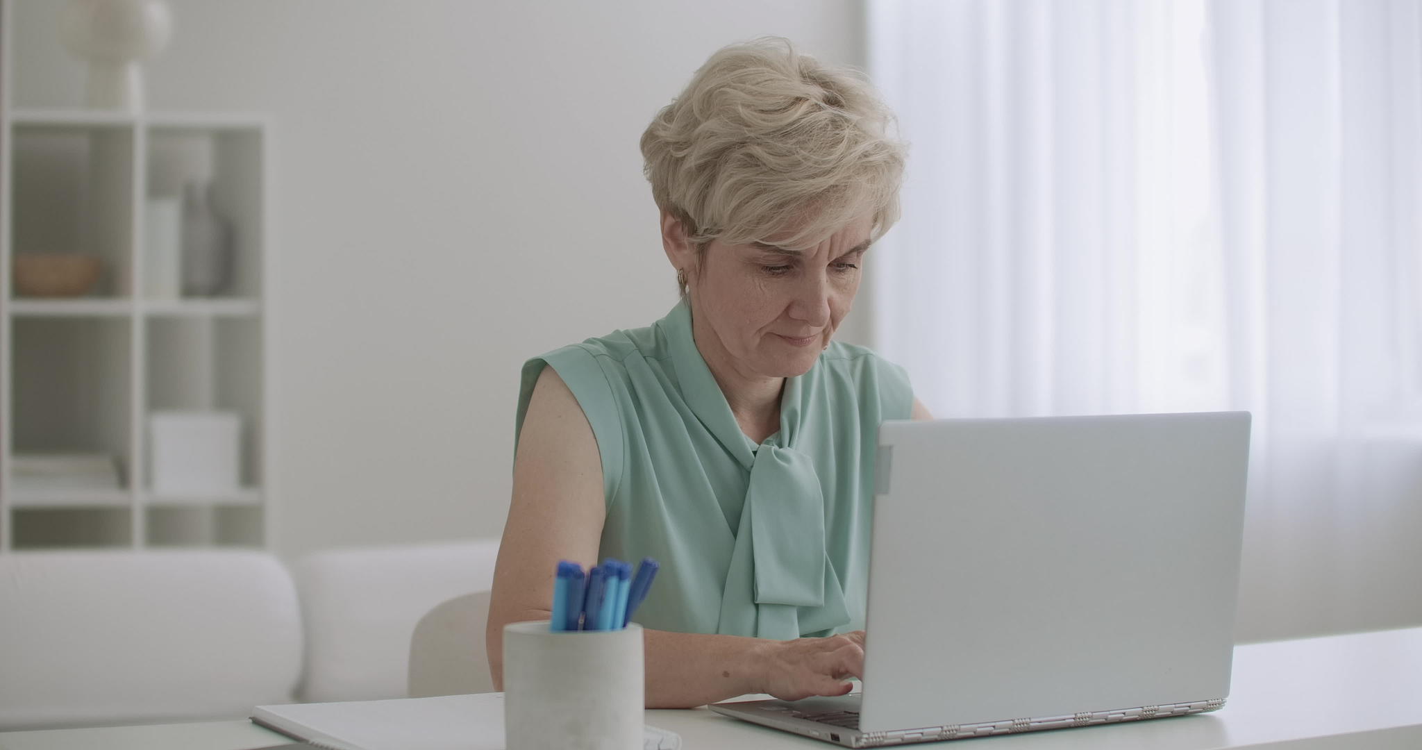 金发老金发女性周末坐在家里的笔记本电脑键盘上视频的预览图