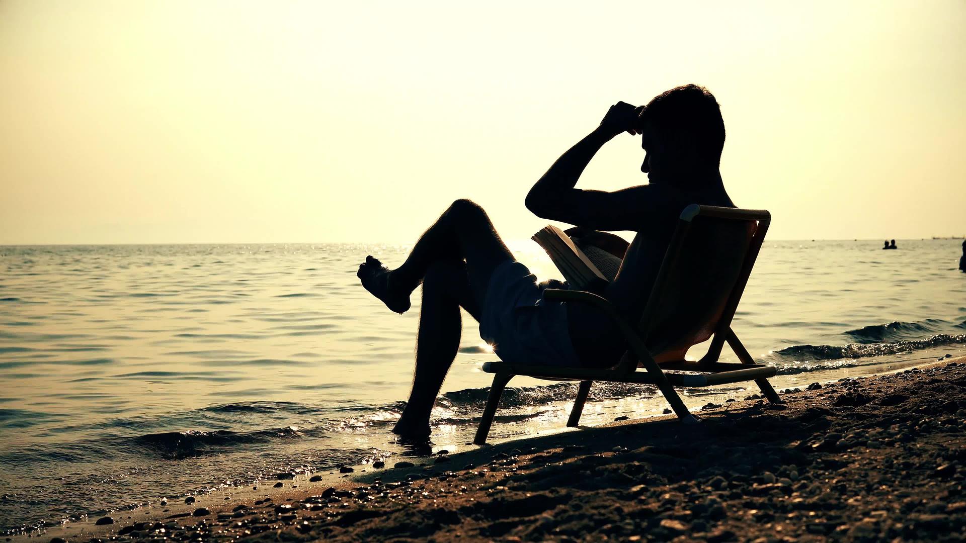 年轻商人坐在浅水中的椅子上读了一本关于海滩日落的书视频的预览图