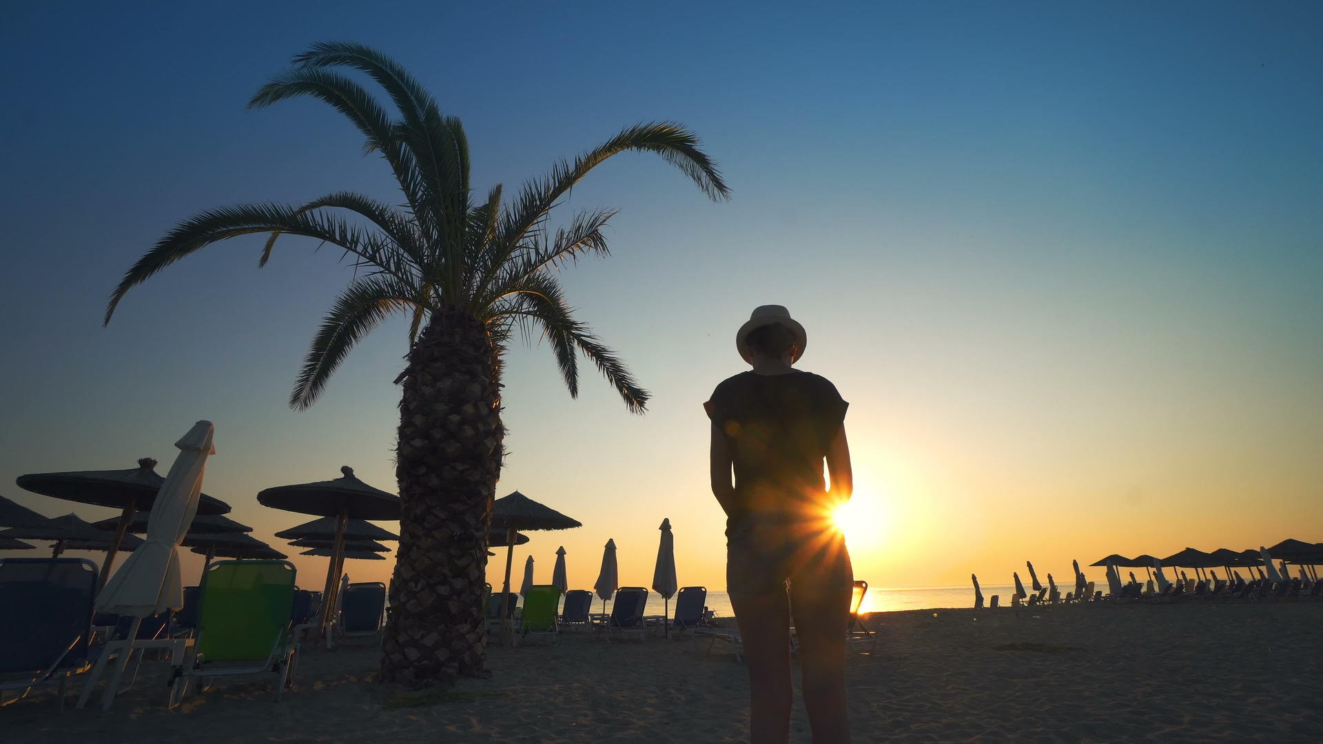 一个女人站在海滩上看着美丽的椰子树、落日棕榈树和雨伞度假胜地视频的预览图