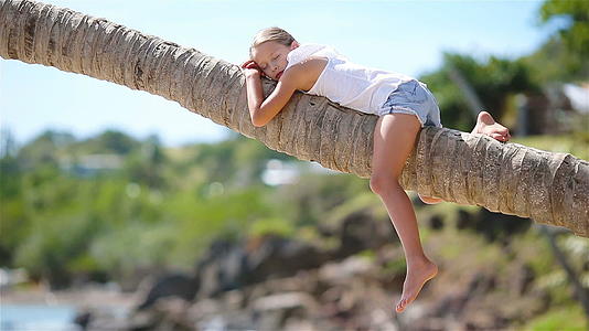 暑假期间小女孩坐在棕榈树上在热带海滩上视频的预览图