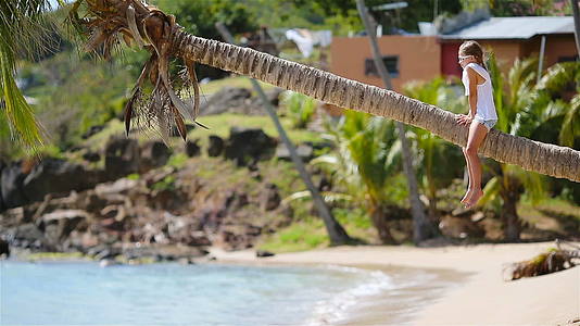 暑假期间可爱的小女孩坐在棕榈树上的热带海滩上视频的预览图