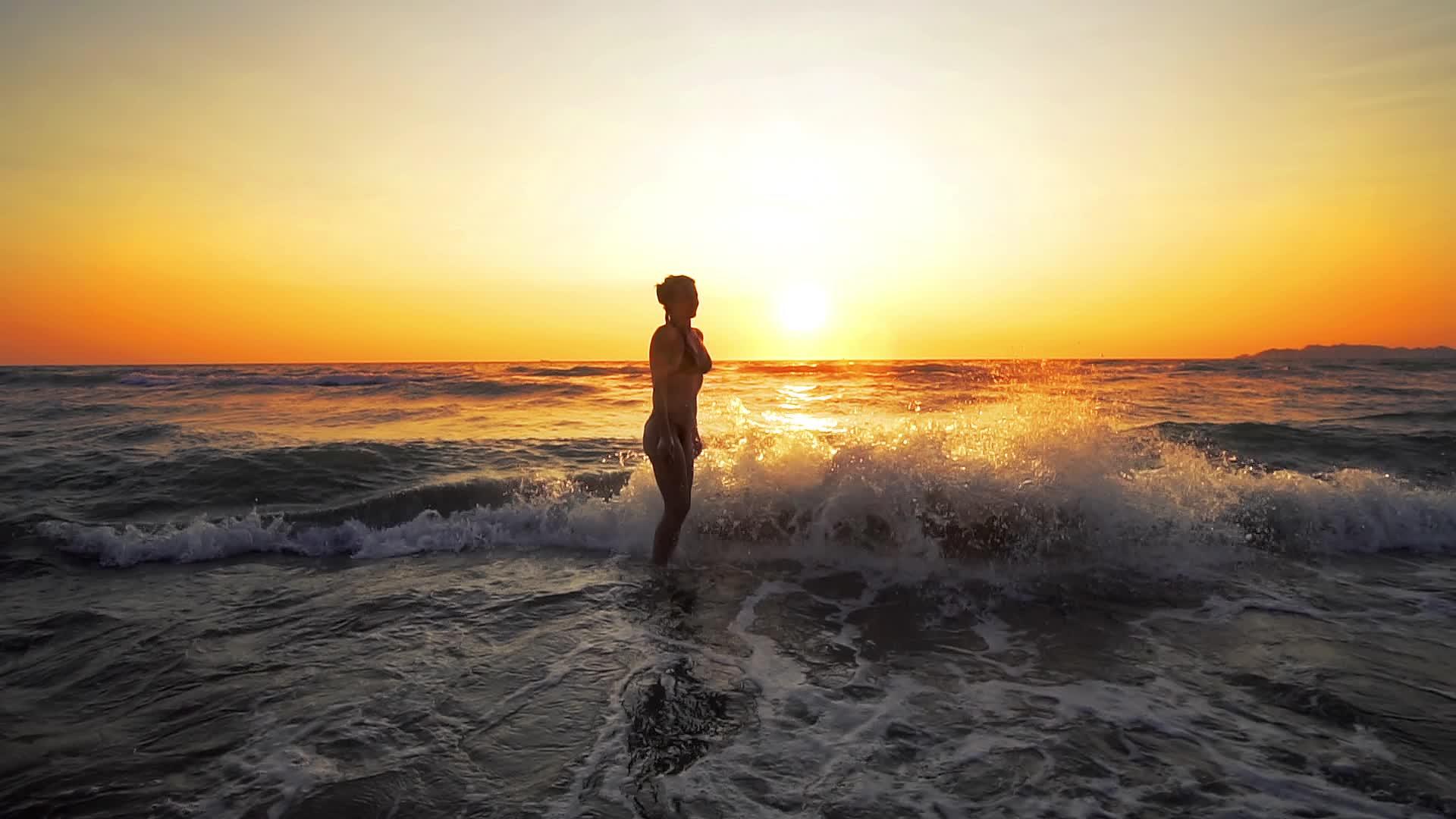 穿着比基尼的年轻女子站在水里日落时被海浪慢慢喷洒视频的预览图