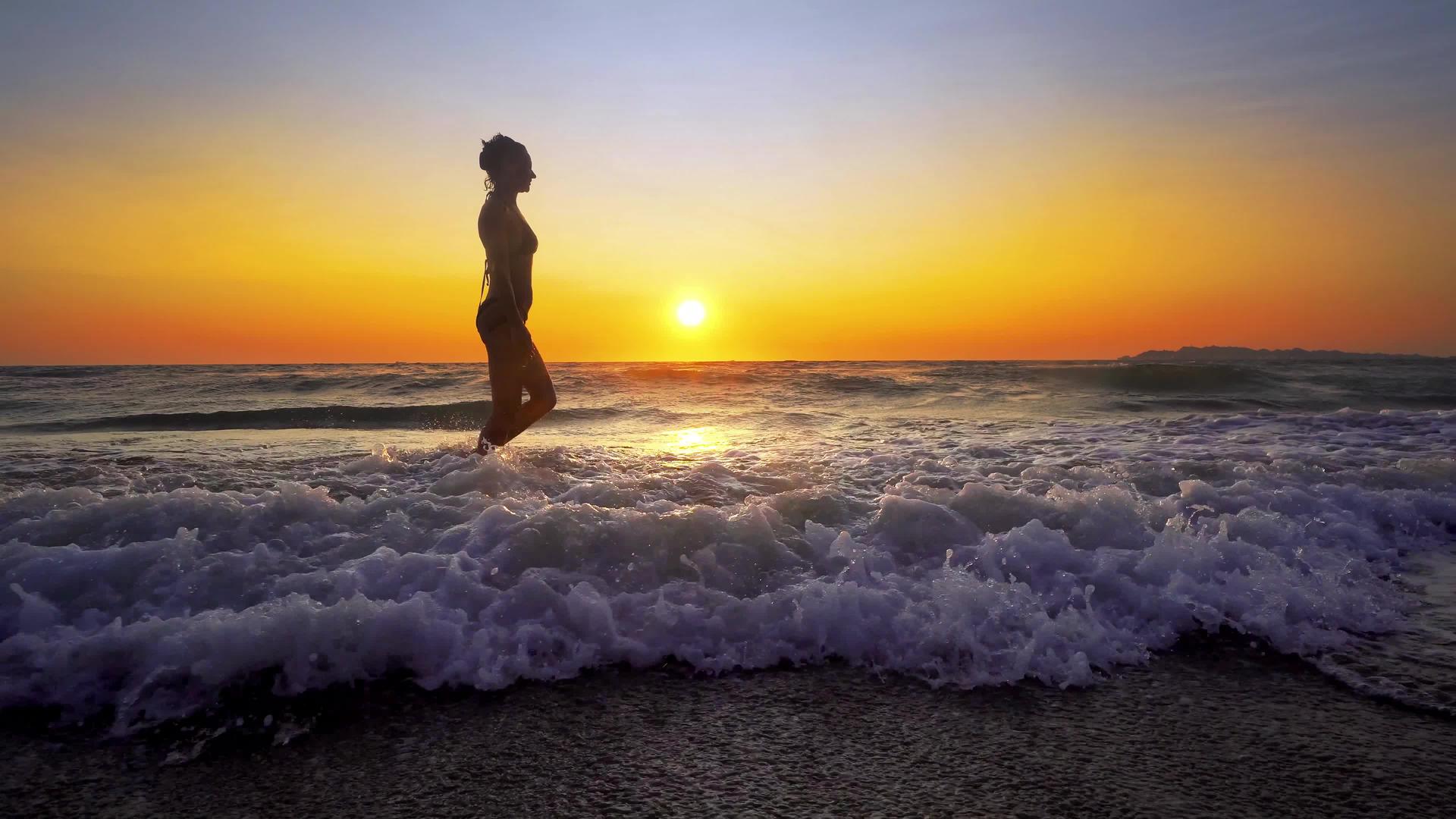 年轻女子在沙滩上散步日落时因海浪喷洒视频的预览图