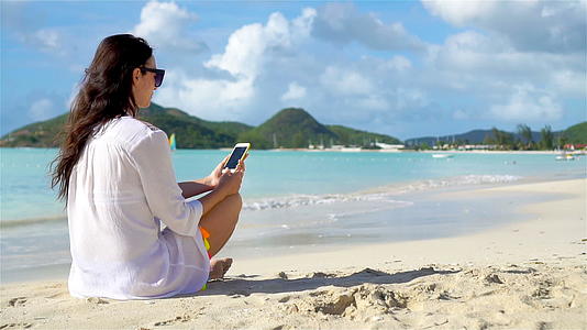 美丽的年轻女孩在热带海滩度假期间使用智能手机视频的预览图