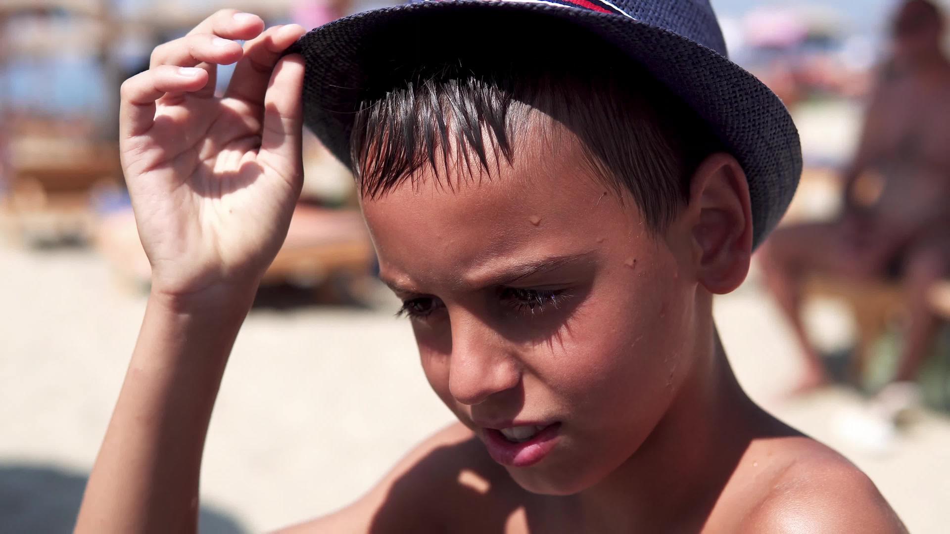 这个戴着帽子的英俊男孩看着相机微笑着坐在海滩上视频的预览图