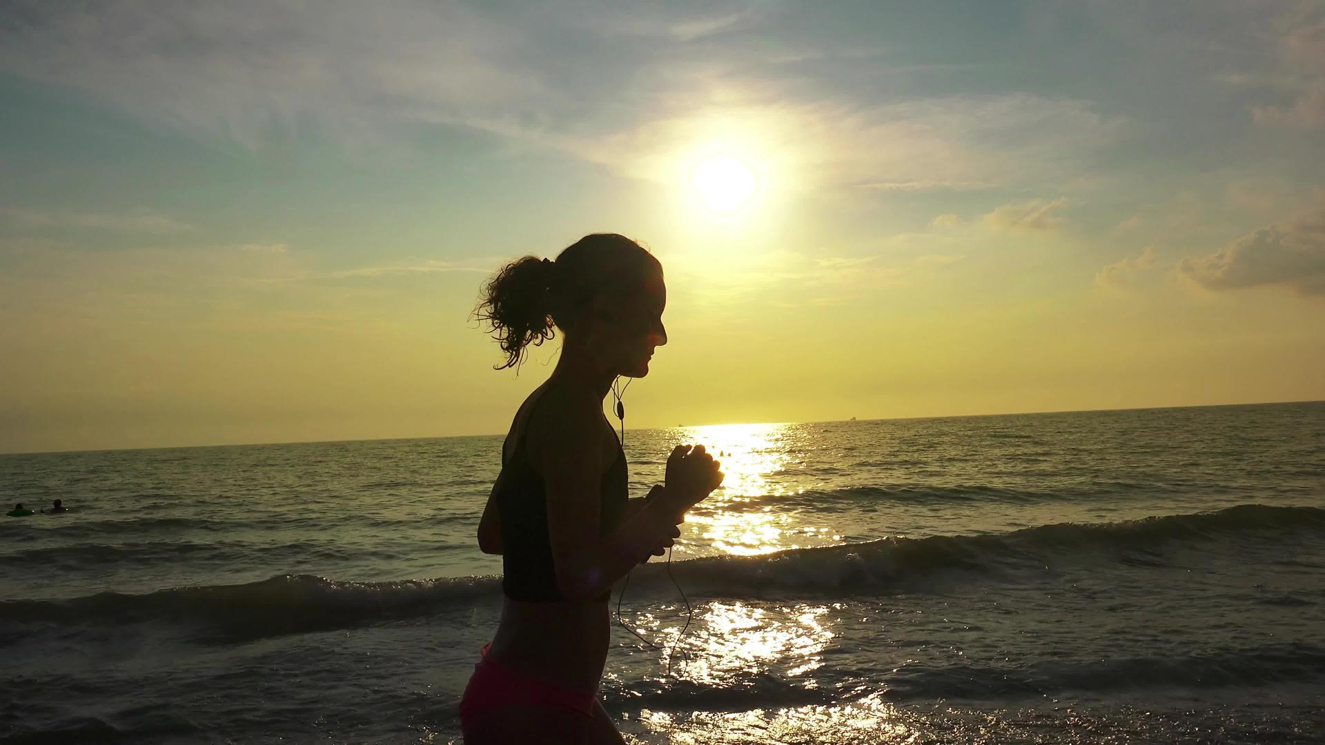 穿着马尾辫和耳机的漂亮女孩在夏天的海滩日落时放松和慢跑视频的预览图