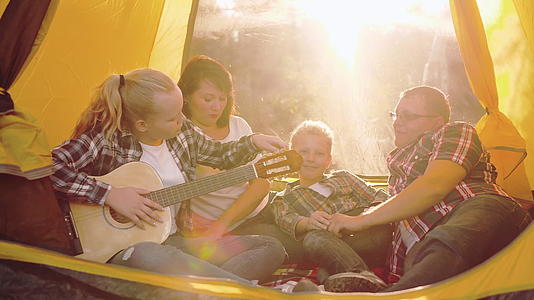 旅游家庭在阳光明媚的森林露营帐篷里休息和演奏吉他音乐视频的预览图