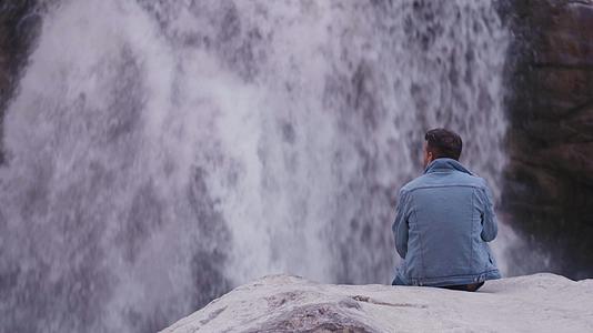 年轻的印度人自信地坐在岩石上穿着蓝色牛仔夹克看着瀑布视频的预览图