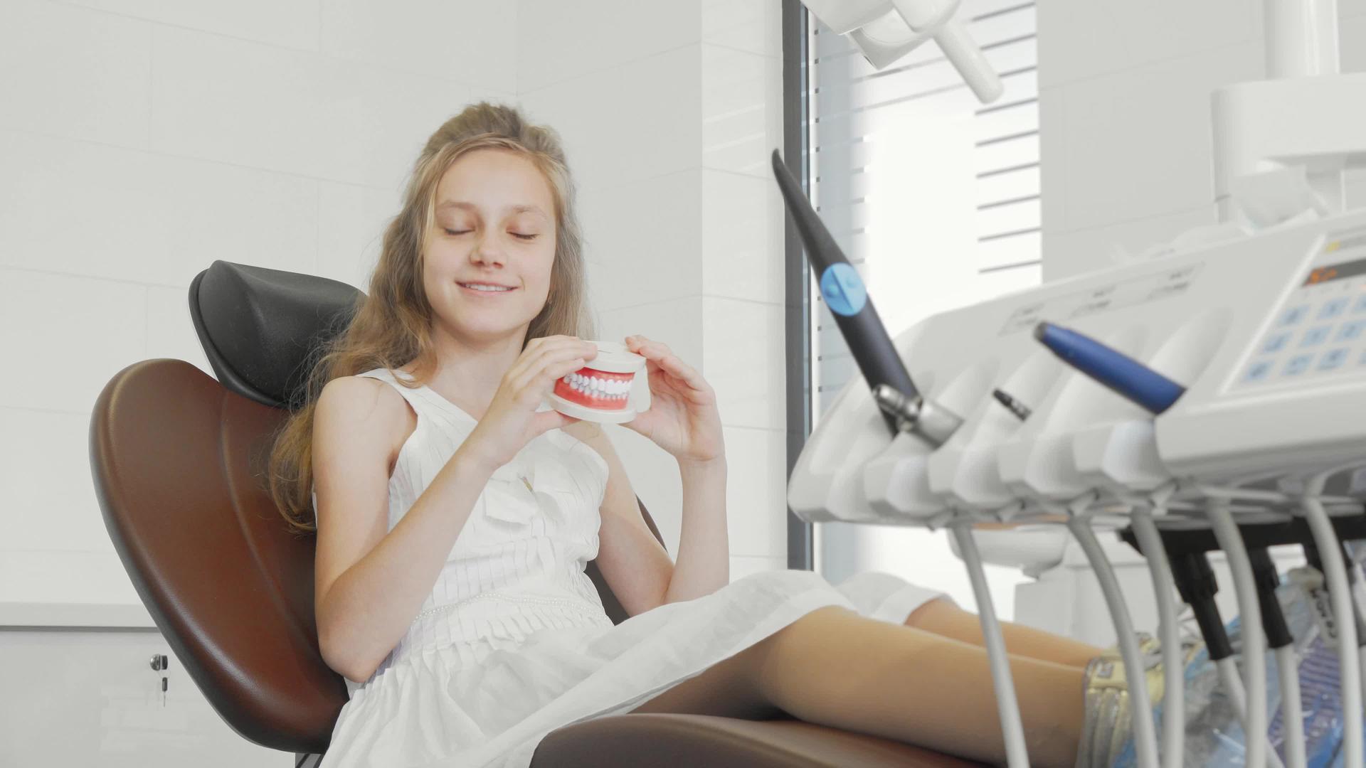 美丽的小女孩坐在诊所的牙科椅子上抱着下巴模型微笑着视频的预览图