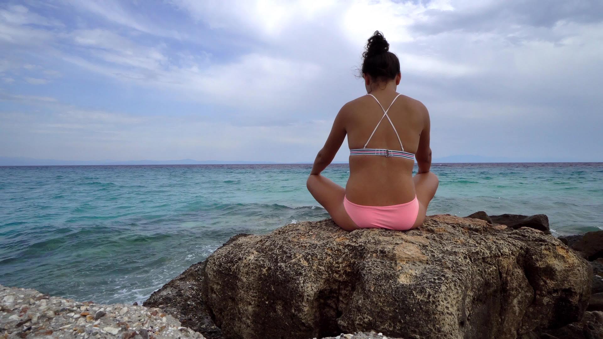 美丽的女人在沙滩上练习瑜伽冥想坐在石头上视频的预览图