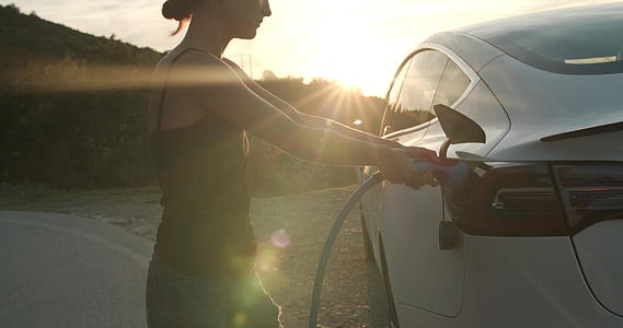 日落时女孩用电动汽车插入充电电缆和手持视频视频的预览图