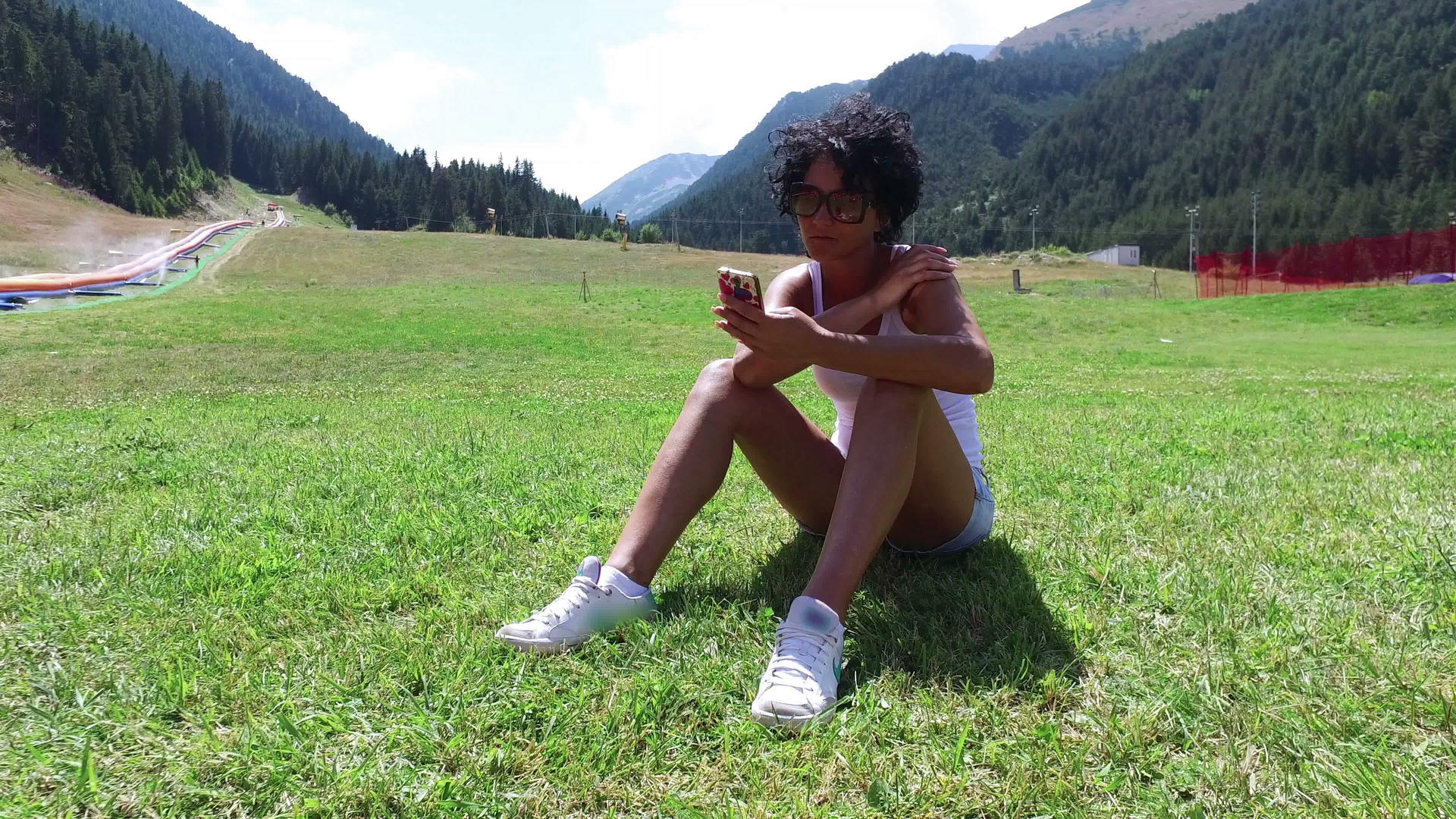 使用智能手机坐在草地上的女性卷发和戴眼镜的女性视频的预览图