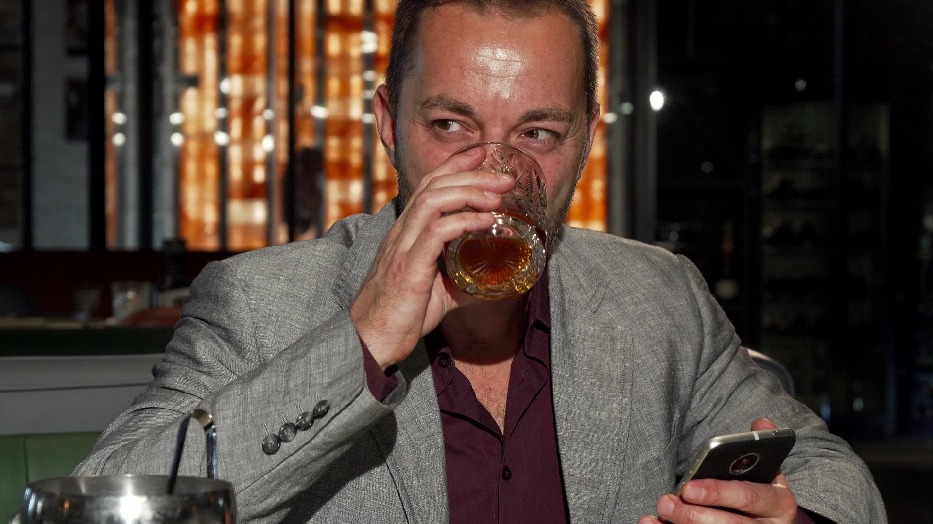 英俊的成熟男子在酒吧喝威士忌时使用智能手机视频的预览图