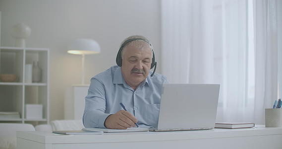 中年男子使用笔记本电脑和耳机通过家中的同事和员工视频的预览图
