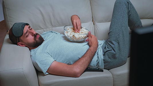 疲惫的男人在沙发上放松在电视机前吃爆米花看电影视频的预览图