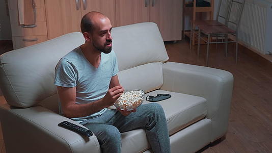 男人手里拿着爆米花碗坐在沙发上同时看电影系列视频的预览图