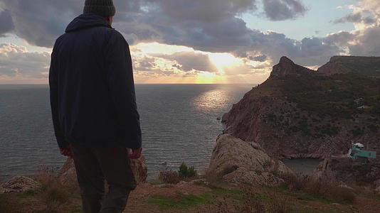 在五颜六色的日落时分穿着夹克和帽子的男人走到海边视频的预览图