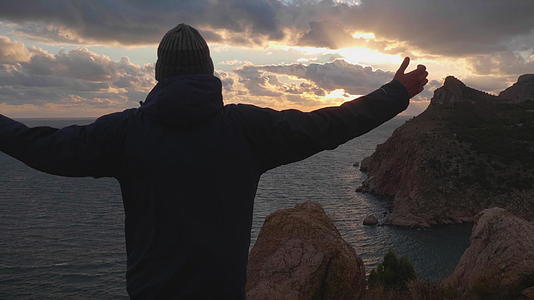 一个穿着夹克和帽子的男人的剪影站在海边岩石悬崖的边缘双手站在海边视频的预览图