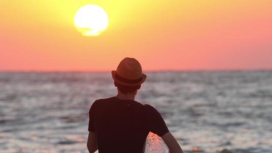 一个戴着充气环帽子的男人看着海边日落的男性旅行者视频的预览图