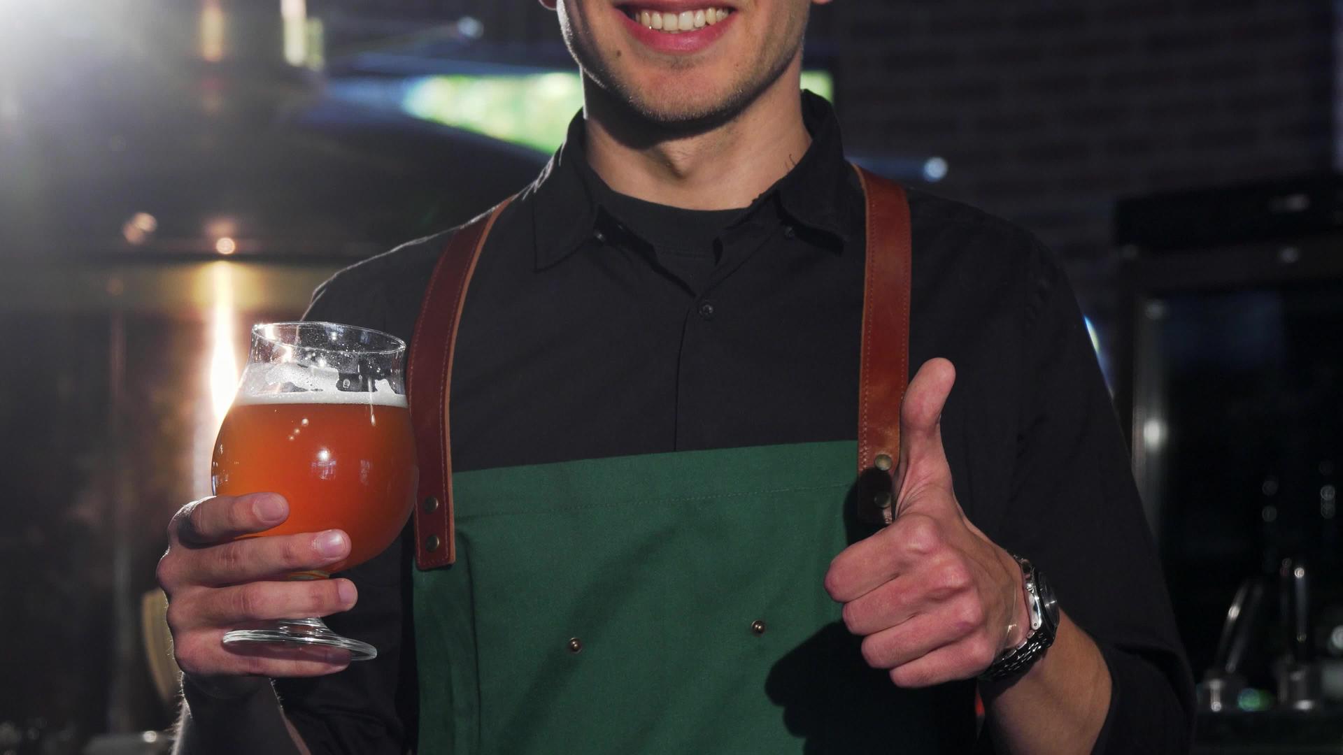 穿着围裙的无法辨认的酿酒师竖起大拇指端着一杯啤酒视频的预览图