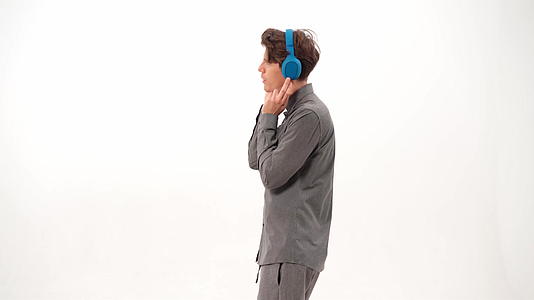 穿着灰色运动服的英俊年轻人戴着蓝色无线耳机跳舞听音乐视频的预览图