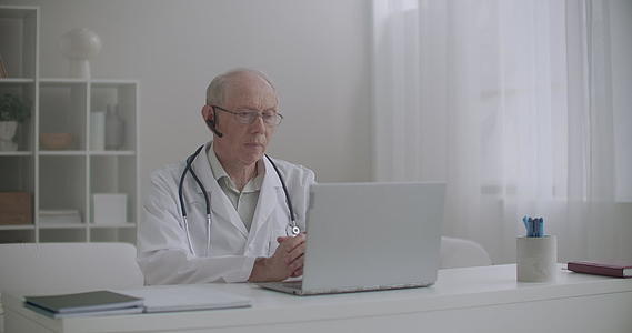 老男医生坐在诊所办公室用笔记本电脑咨询病人视频的预览图