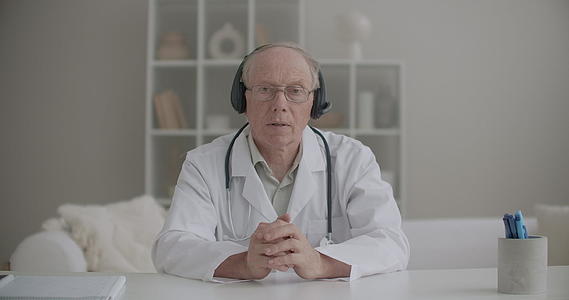 穿着白色长袍眼镜和耳机的男医生正在在线拍摄正面媒体肖像视频的预览图