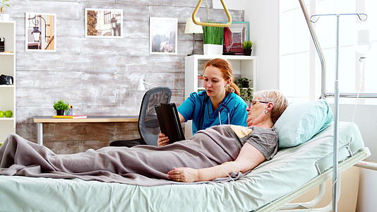 女护士帮助躺在医院床上的老年残疾妇女使用数字平板电脑视频的预览图