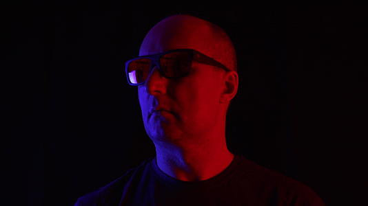 在蓝色和红色光线的黑色背景上戴太阳镜的秃头男人在黑暗中视频的预览图