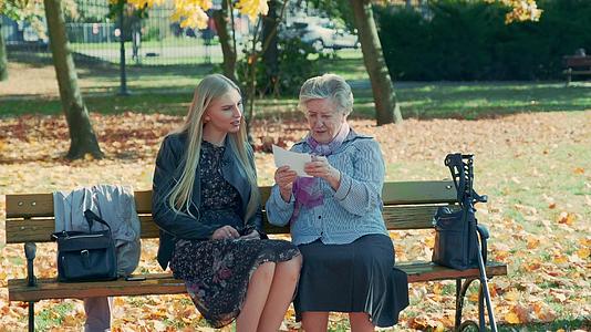 年轻的金发女郎听着老妇人坐在长椅上看东西视频的预览图