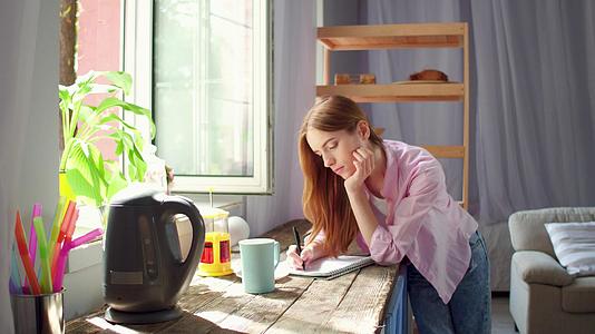 站在厨房木桌旁的年轻女子喝茶做笔记视频的预览图