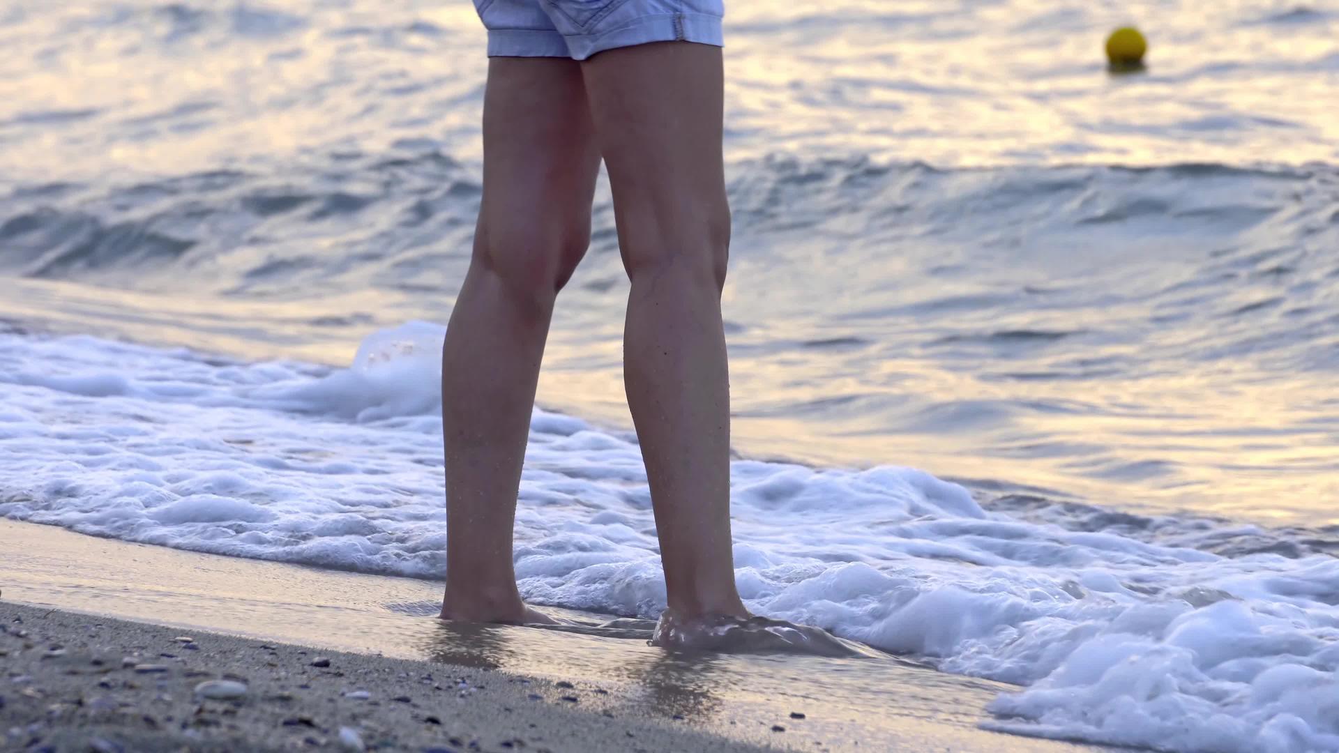 站着赤脚的女人在夏天的海滩上玩海浪喷她的腿视频的预览图