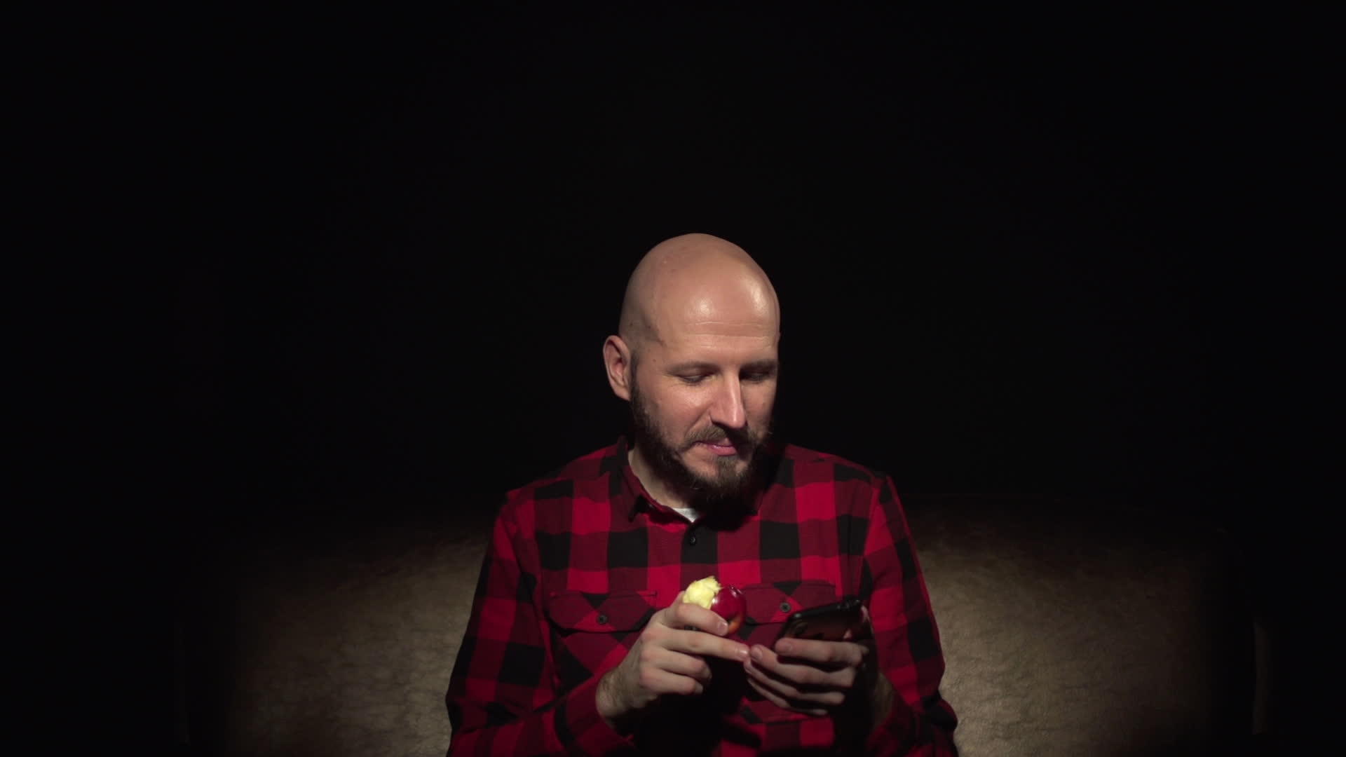 胡子穿着红色格子衬衫吃着苹果在智能手机上翻滚有趣而有趣视频的预览图