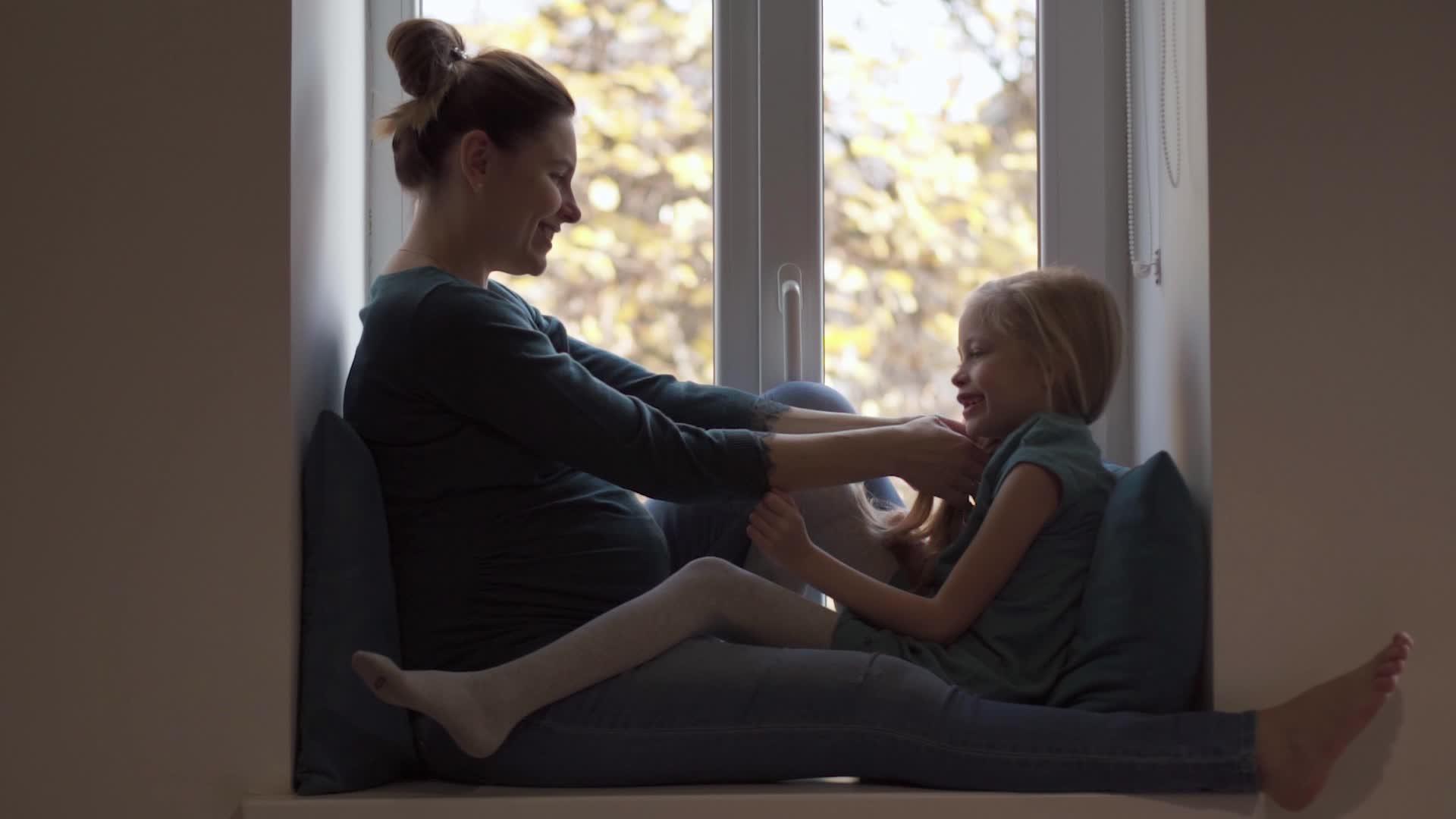 穿着绿色衣服的孕妇和女儿在窗台上互相抚摸视频的预览图