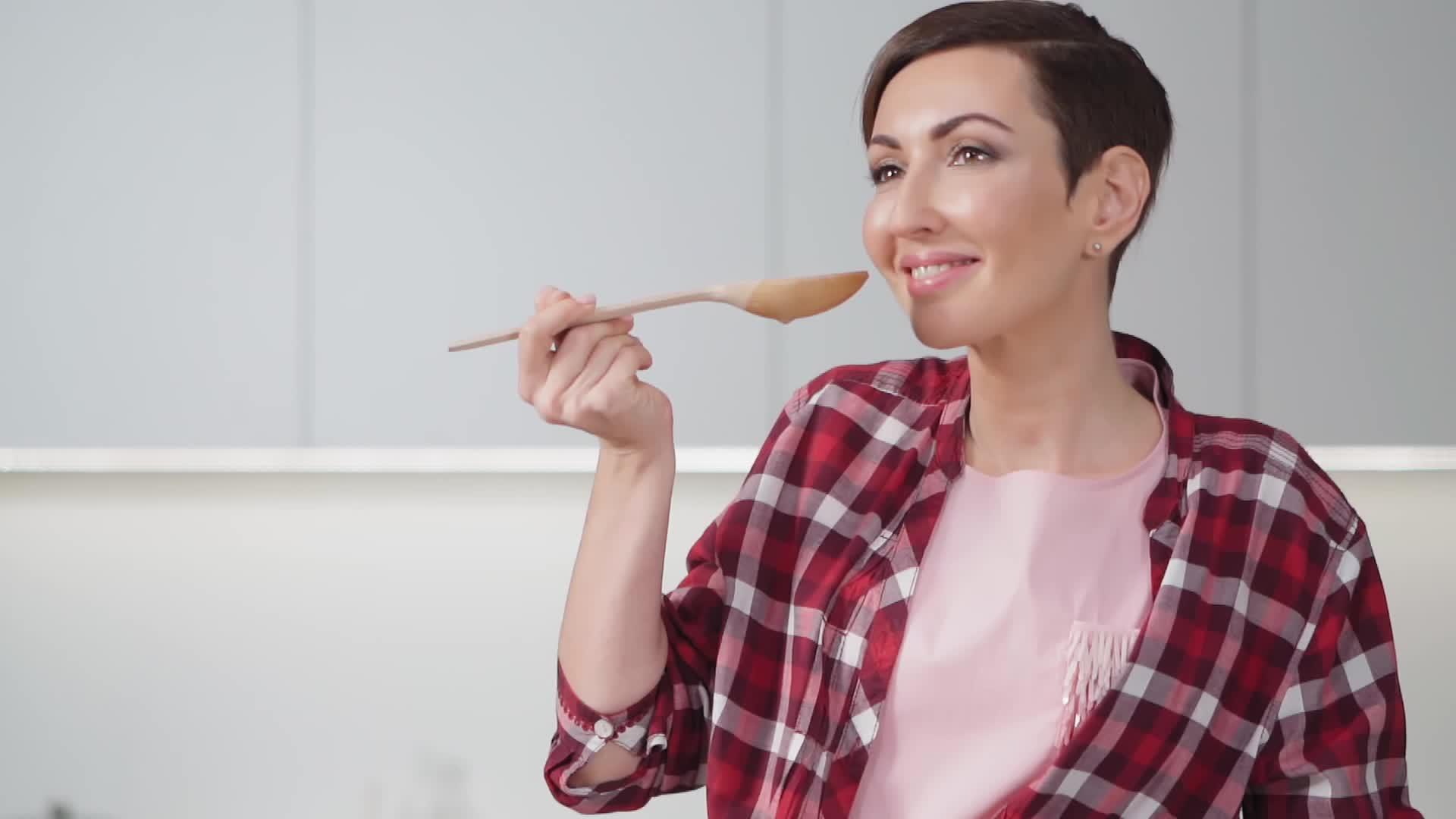 美丽的家庭主妇用长木勺用短发做饭试着站在现代厨房里视频的预览图
