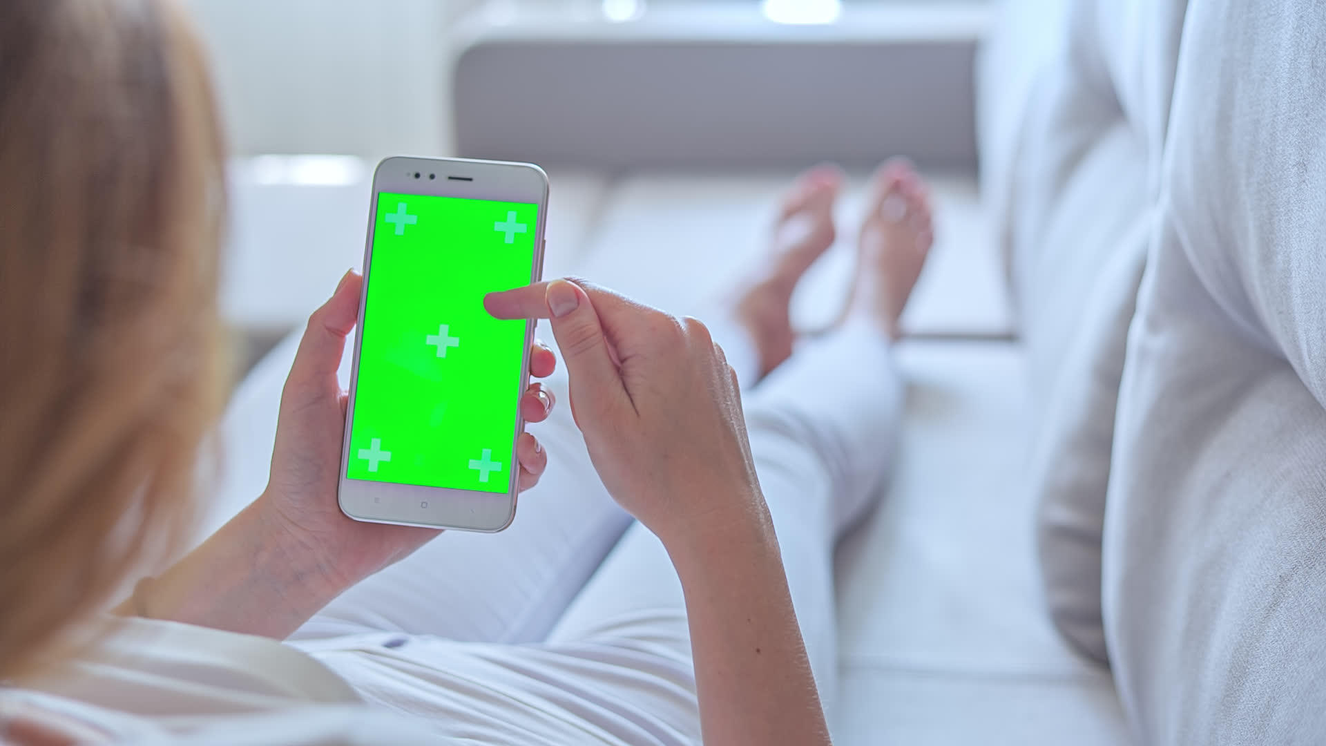 穿着白色牛仔裤的年轻女性躺在沙发上在绿色屏幕上使用智能手机视频的预览图