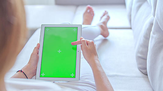 穿着白色牛仔裤的年轻女性躺在沙发上使用绿色屏幕平板电脑视频的预览图