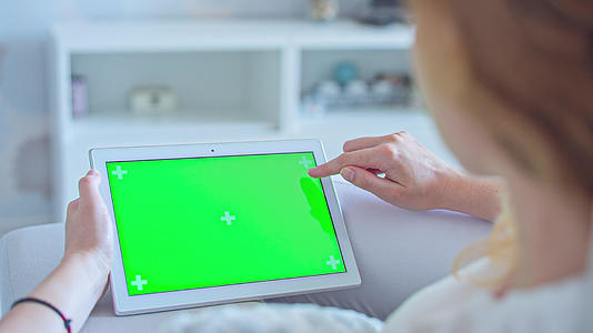 穿着白色牛仔裤的年轻女性躺在沙发上使用绿色屏幕平板电脑视频的预览图