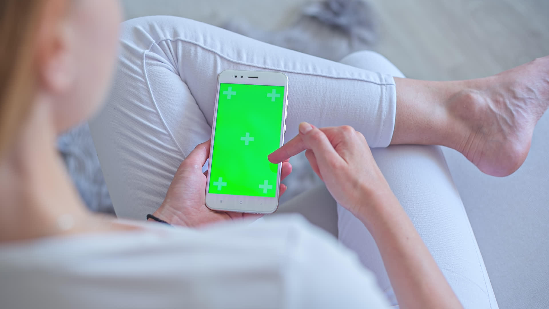 年轻女性穿着白色牛仔裤坐在沙发上使用智能手机和绿色屏幕视频的预览图