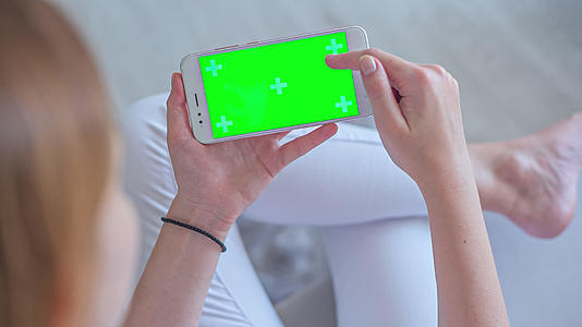 年轻女性穿着白色牛仔裤坐在沙发上使用智能手机和绿色屏幕视频的预览图