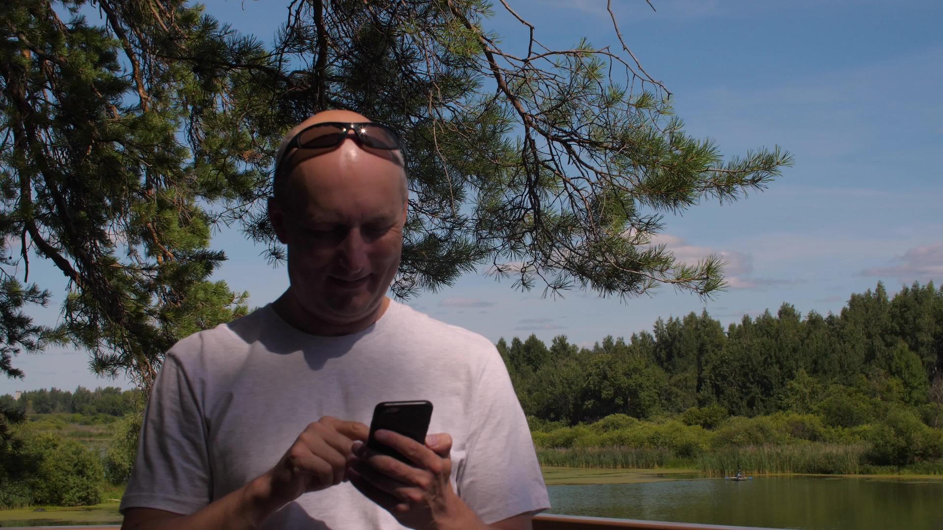 英俊的男人在树上和池塘景观的手机上打字视频的预览图