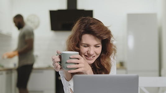 微笑的白女早上在厨房的笔记本电脑上读好消息视频的预览图