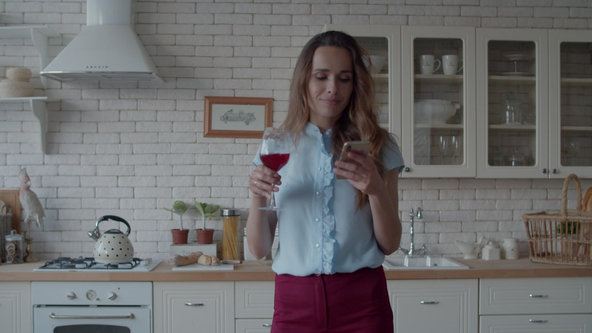 迷人的女孩拿着酒杯在豪华厨房看手机视频的预览图