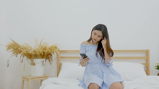漂亮的女人戴着耳机坐在床上在手机应用中听音乐视频的预览图