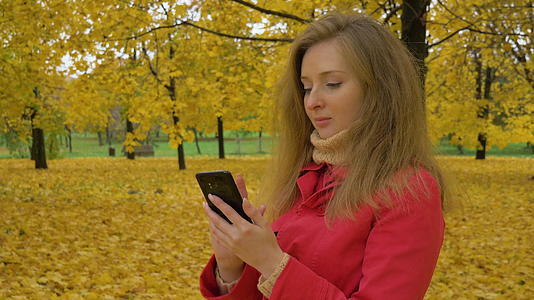 秋天穿着红色斗篷的年轻美女在公园的黄色树林里用手机打字视频的预览图