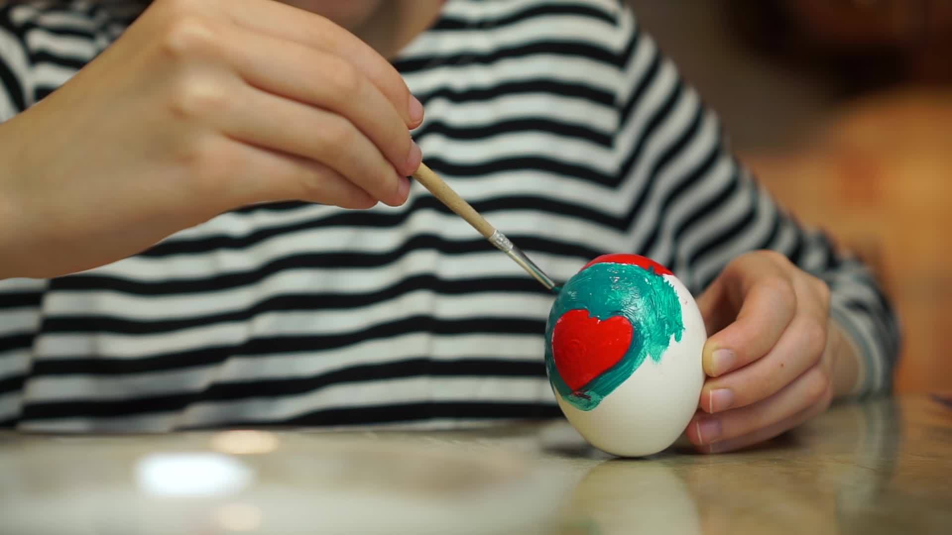 条纹礼服中的特写儿童和女孩手绘复活节彩蛋和厨房桌子上的油漆以及红色的心形视频的预览图